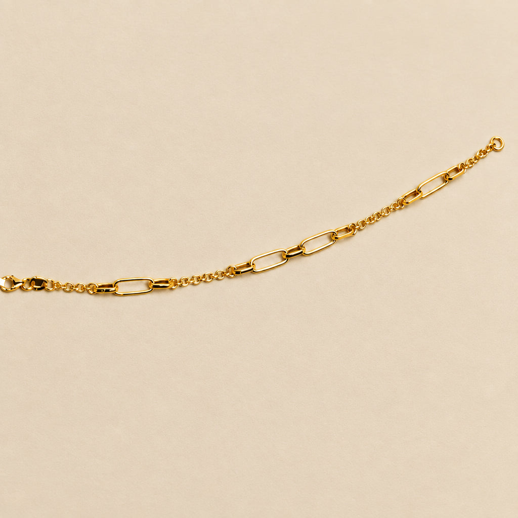 A gold paperclip chai bracelet laid horizontally against a plain beige background.