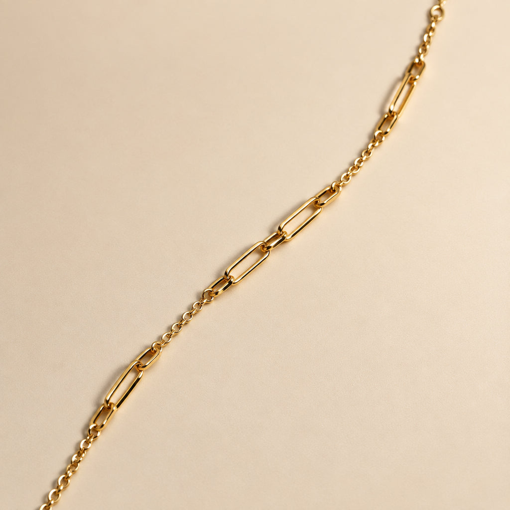 A gold paperclip chain bracelet laid diagonally on a plain beige background.