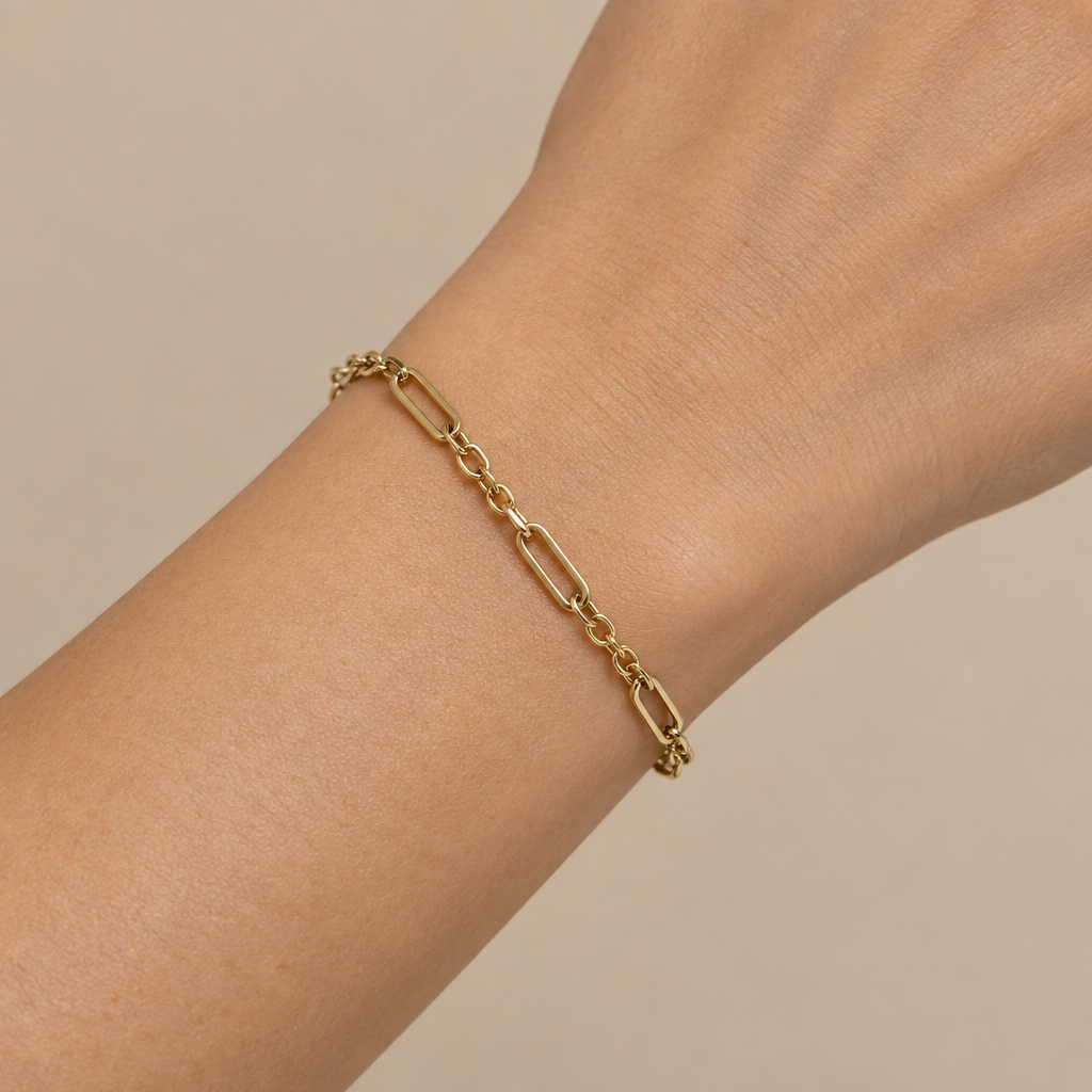 A delicate gold paperclip chain bracelet worn on a person's wrist, shown against a soft beige background.